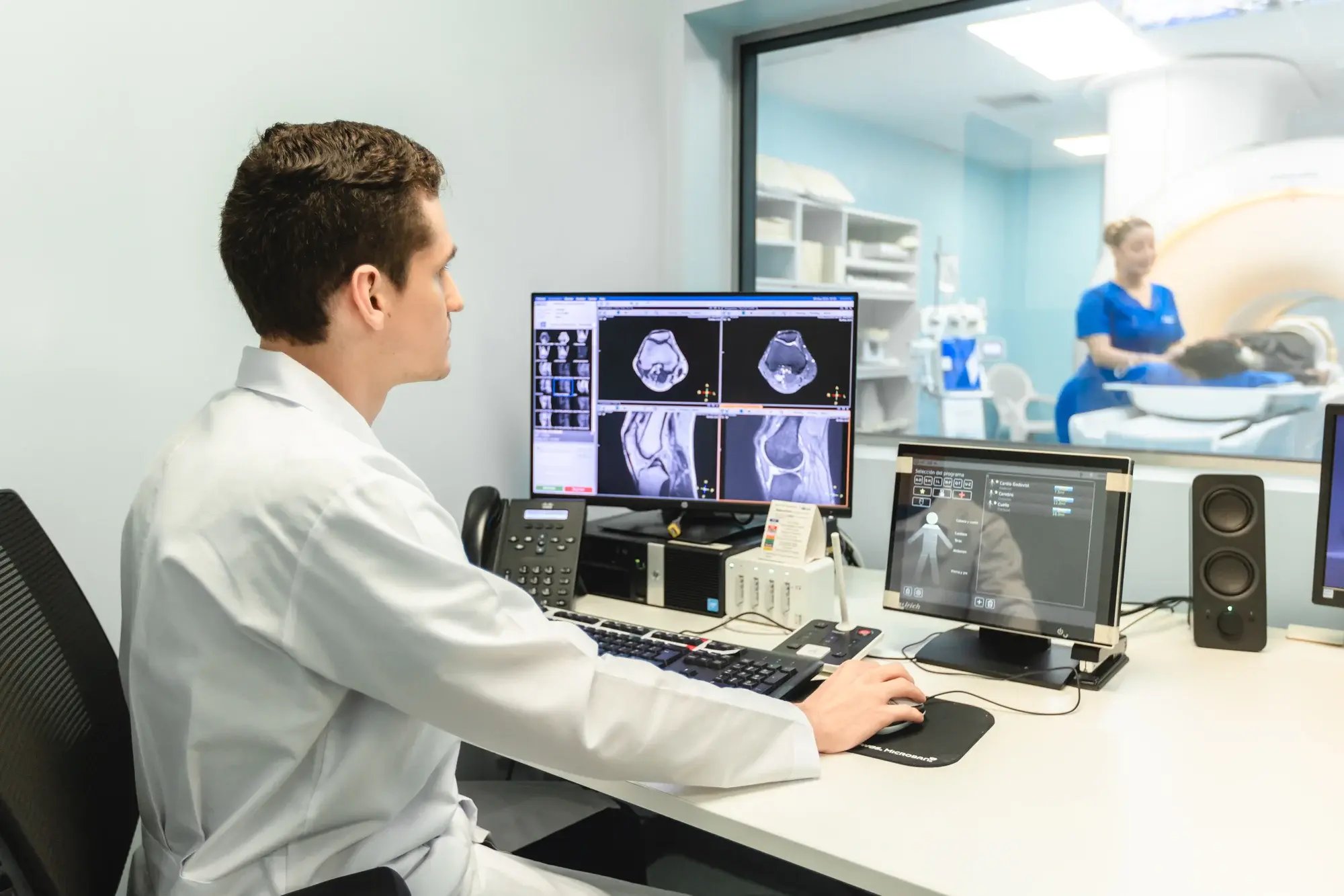Radiólogo revisando imágenes de resonancia magnética en monitores, paciente en máquina de resonancia al fondo.
