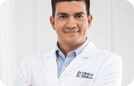 Médico con bata blanca de Clínica Bíblica sonriendo frente a cámara.