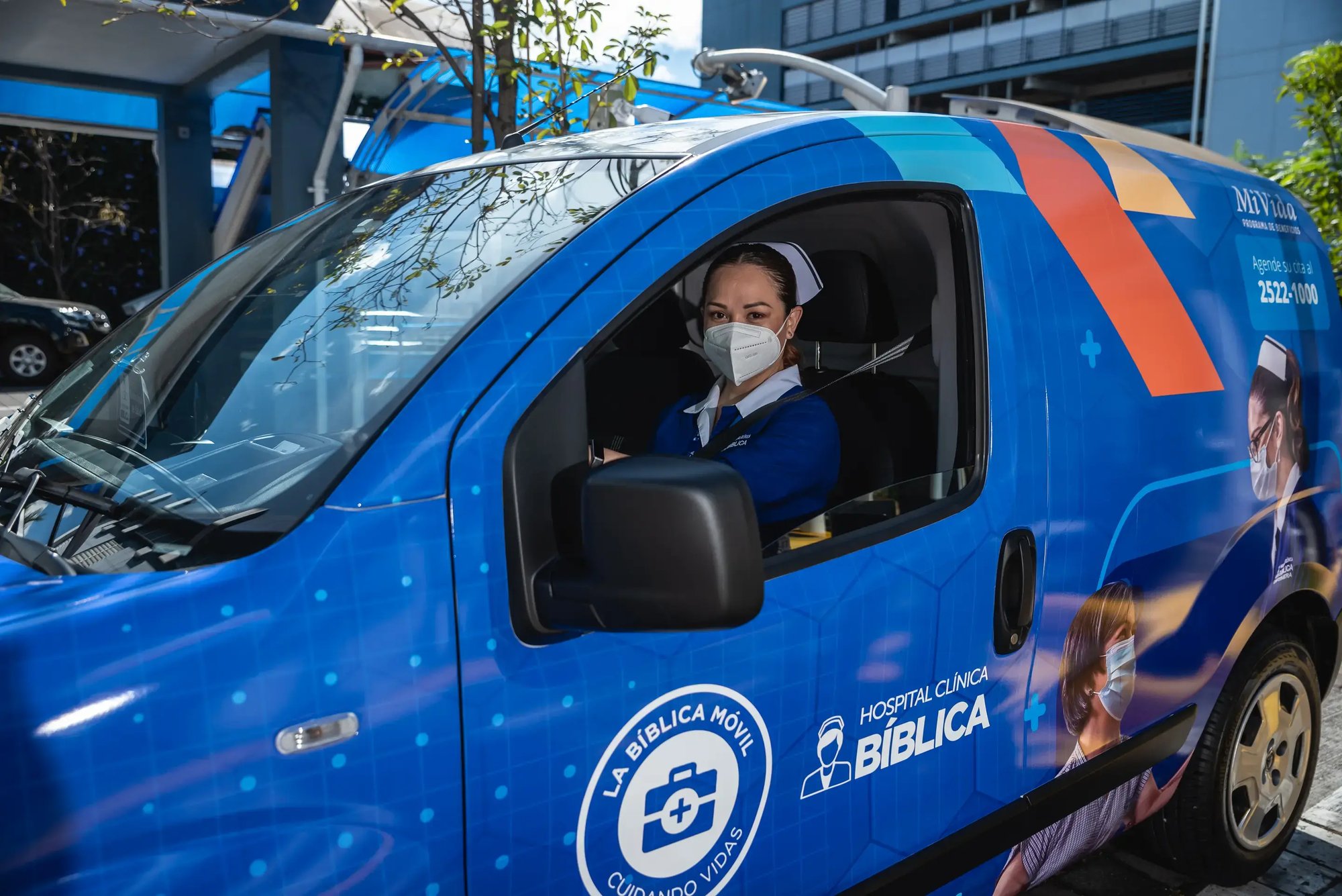 Nurse from Clínica Bíblica sitting behind the wheel of a Bíblica Móvil unit, wearing a mask and blue uniform.