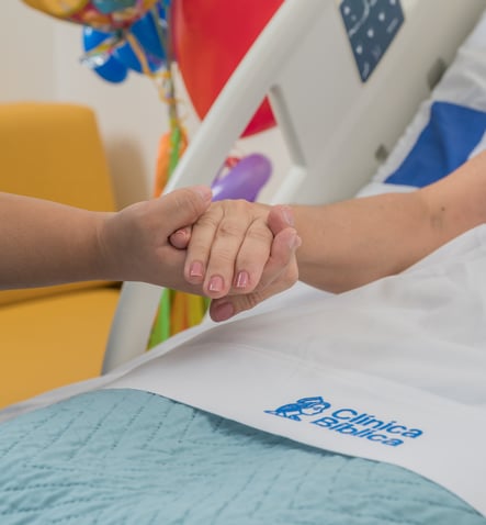 Manos de una persona sosteniendo la mano de una paciente en cama con manta azul y logotipo de Clínica Bíblica. Manos de una persona sosteniendo la mano de una paciente en cama con manta azul y logotipo de Clínica Bíblica.
