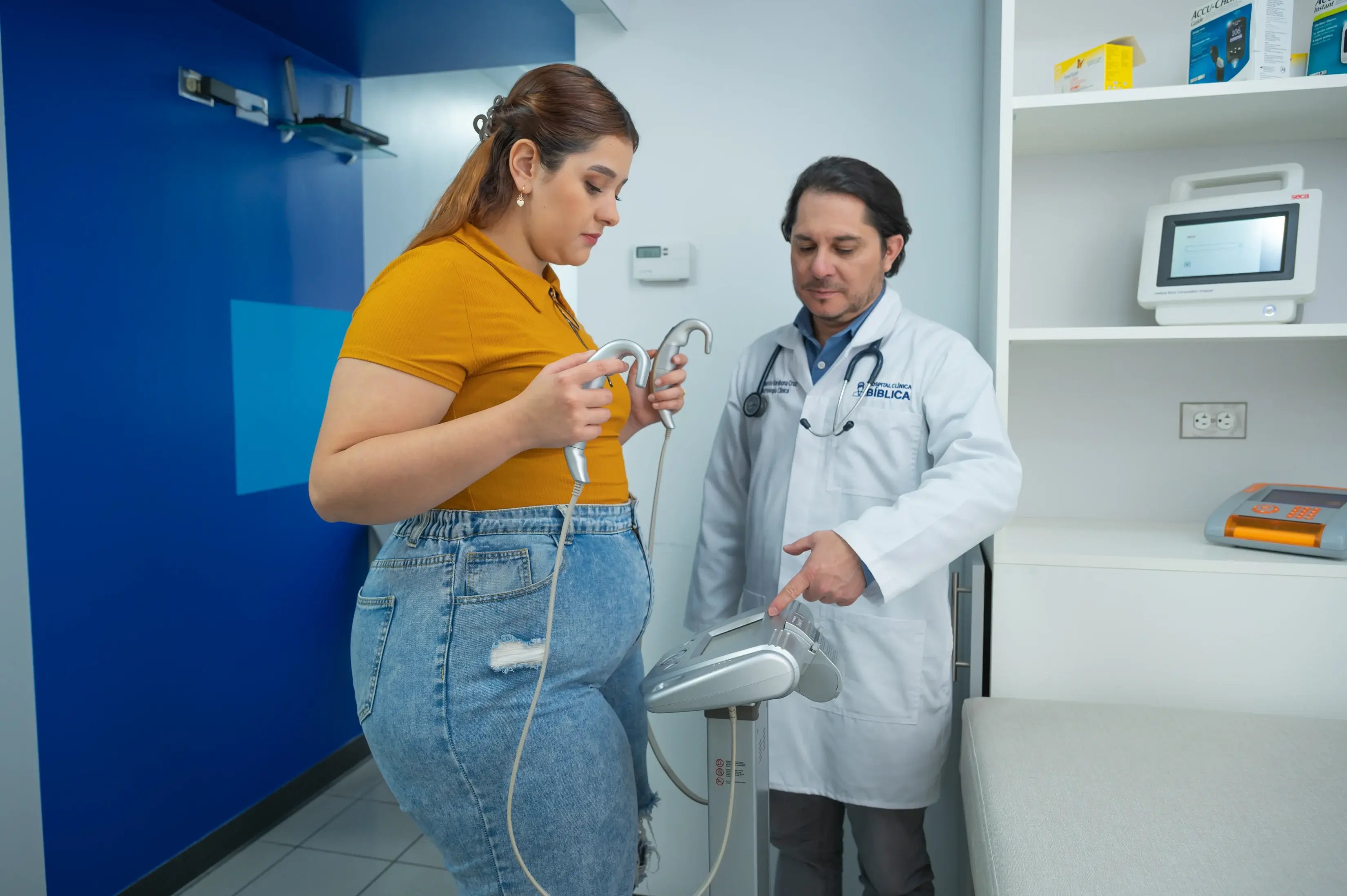 Evaluación clínica con médico revisando equipo de bioimpedancia mientras paciente sostiene manilares.