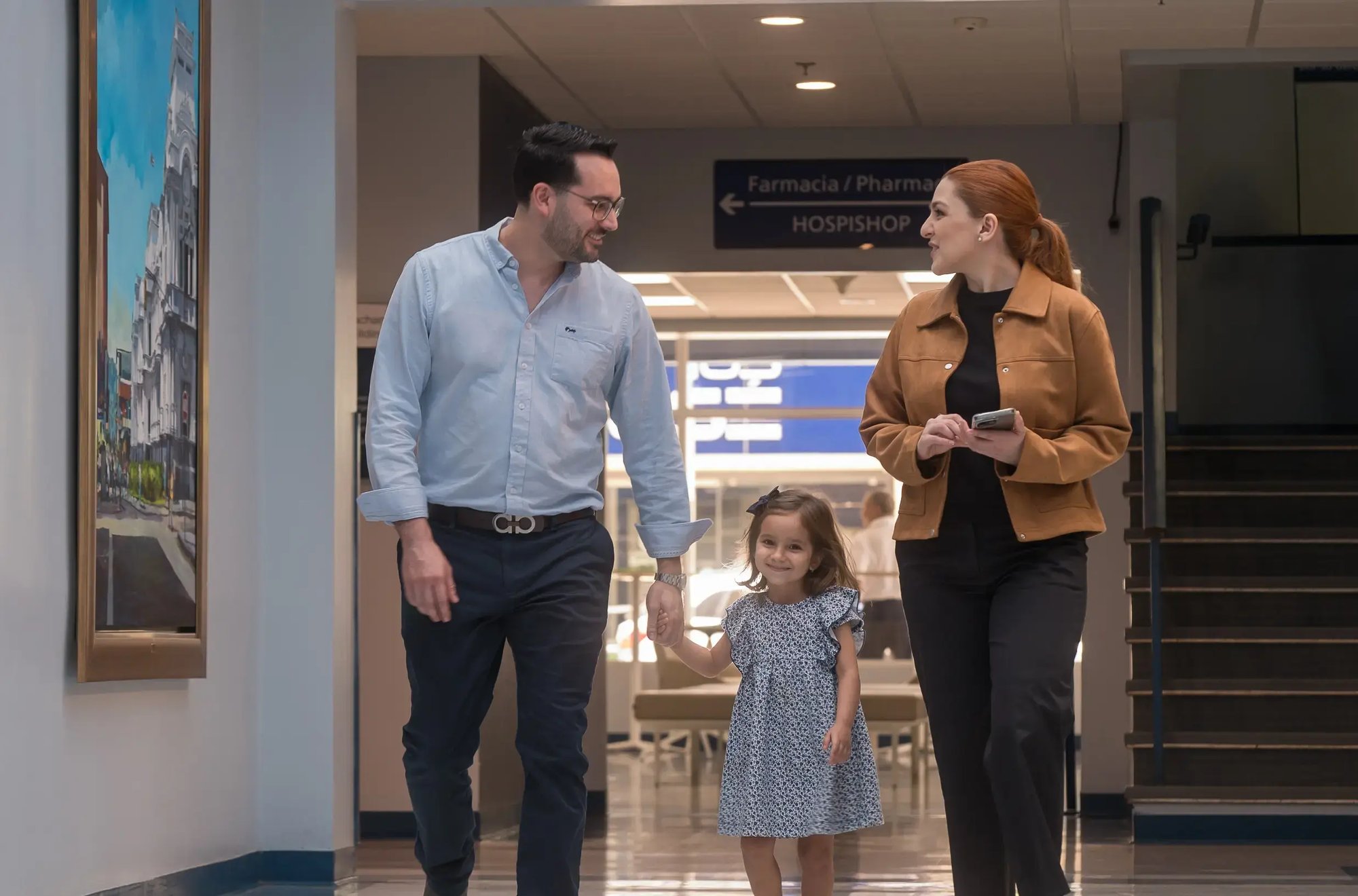 Familia caminando por un pasillo de la clínica junto a carteles de farmacia y servicios.