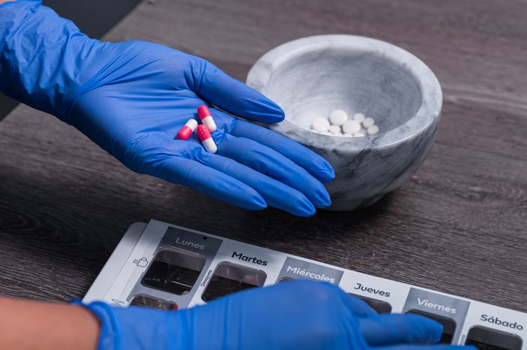 Mano con guantes azules sosteniendo cápsulas rojas y blancas junto a un mortero con tabletas y un organizador semanal de medicamentos.