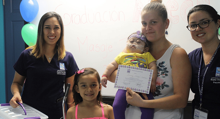 Grupo de mujeres y niñas posando en un aula durante una actividad de graduación, una mujer sostiene un certificado y un bebé.