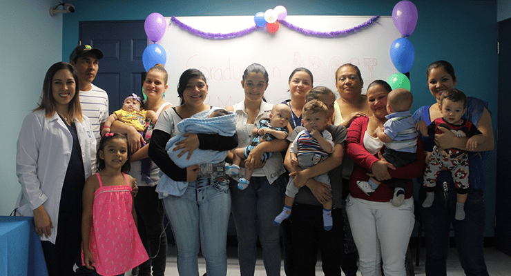 Grupo de adultos y bebés posando frente a un pizarrón decorado con globos durante una actividad grupal en un aula.