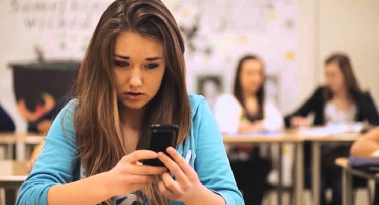 Adolescente sentada en un aula mirando un teléfono móvil con otras personas al fondo