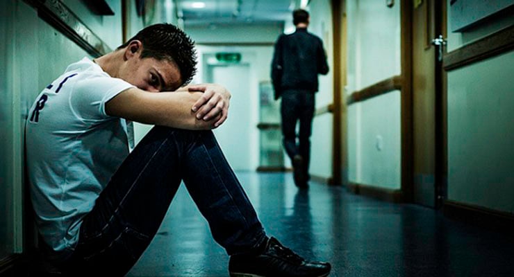 Adolescente sentado en el suelo de un pasillo escolar, con la cabeza apoyada en los brazos y otra persona caminando al fondo del pasillo.