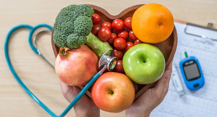 Manos sosteniendo un recipiente en forma de corazón con frutas y vegetales frescos junto a un glucómetro y estetoscopio sobre una mesa