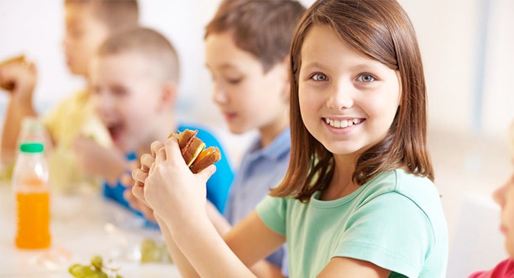 Niña sonriendo mientras come un sándwich junto a otros niños en una mesa