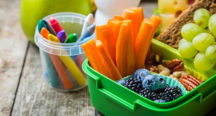 Lonchera escolar verde con zanahorias, arándanos, uvas y nueces junto a un recipiente con crayones de colores sobre una mesa de madera.