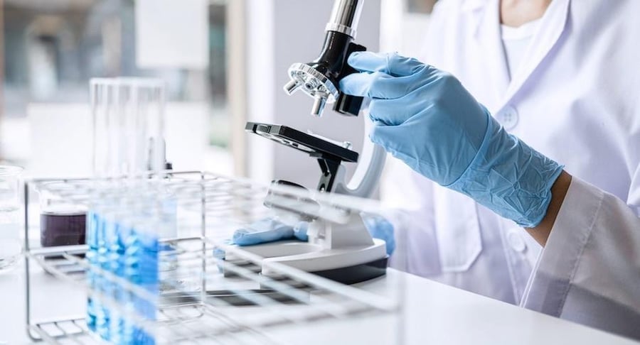 Profesional de laboratorio con guantes azules ajustando un microscopio cerca de gradillas con tubos de ensayo que contienen muestras líquidas.
