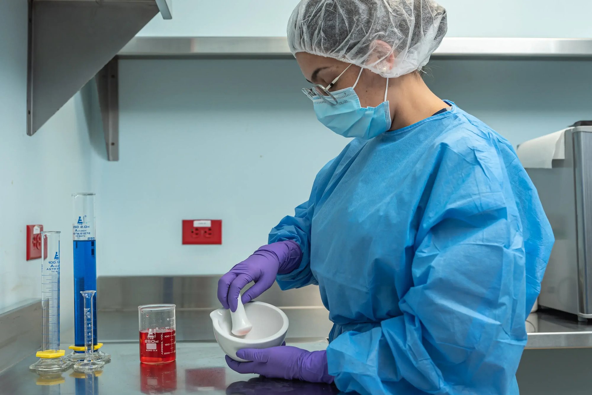 Laboratory analyst preparing sample in mortar next to beakers with colored liquids.