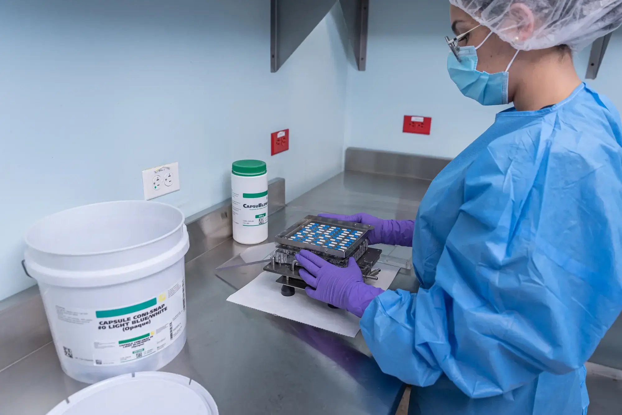 Técnica farmacéutica manipulando bandeja con cápsulas en proceso de control de calidad.