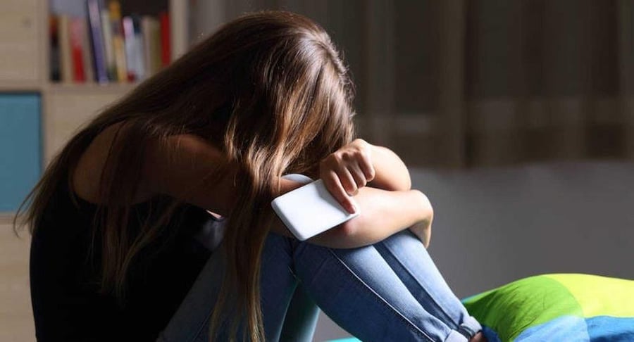 Joven sentada en su cama en una habitación oscura, abrazando sus rodillas y sosteniendo un celular, representando sentimientos de ansiedad y preocupación.
