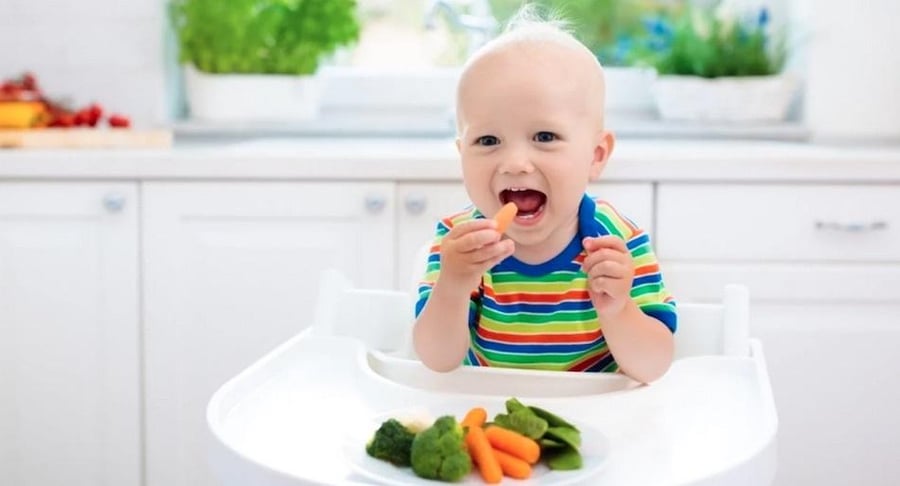 Bebé sonriente sentado en una silla alta de comer mientras sostiene una pequeña zanahoria y tiene un plato con brócoli y vegetales frente a él.