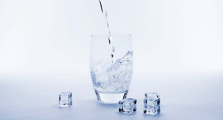 Agua cristalina vertiéndose en un vaso de vidrio transparente rodeado de cubos de hielo sobre una superficie blanca brillante.
