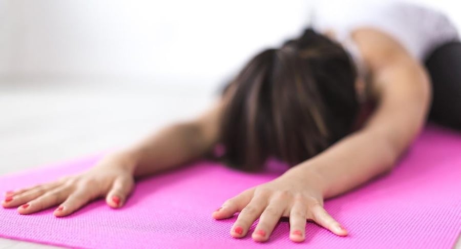Una mujer con uñas rojas realiza un estiramiento de espalda extendiendo sus brazos sobre un mat de yoga rosado en el suelo.