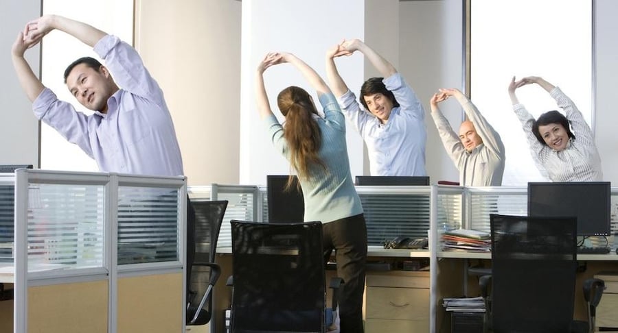 Grupo de compañeros de oficina de pie en sus cubículos realizando estiramientos de brazos y espalda como parte de una rutina de pausas activas.