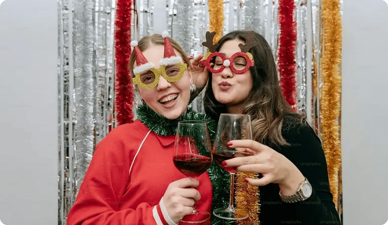 Dos personas con lentes navideños sosteniendo copas de vino tinto frente a un fondo decorado con tiras brillantes.