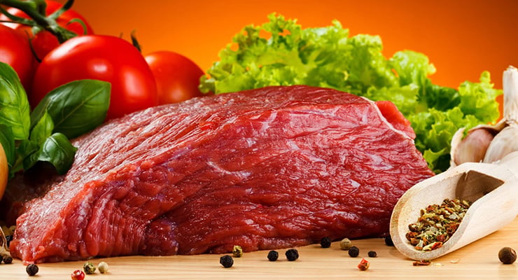 Corte de carne cruda sobre tabla de madera acompañado de tomates, lechuga, ajo y especias.
