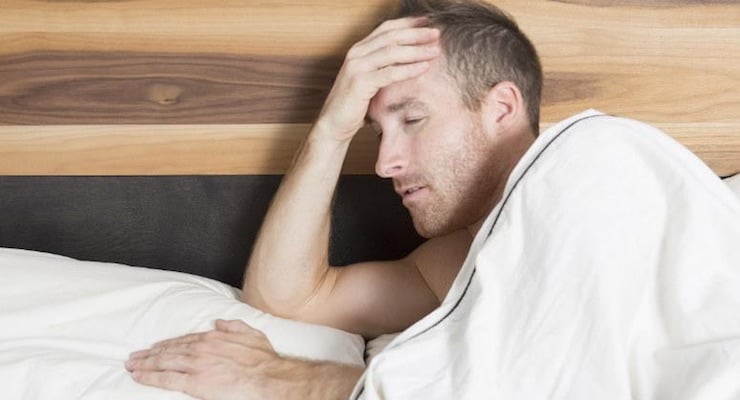 Un hombre joven sentado en una cama, con una mano en la frente y expresión de preocupación o frustración, ilustrando problemas de salud sexual masculina.