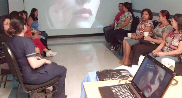 Grupo de mujeres participando en una charla educativa con proyección audiovisual en una sala.