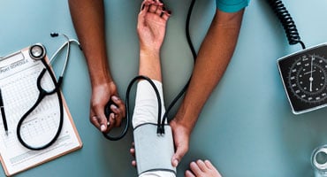 Vista aérea de un médico tomando la presión arterial a un paciente, rodeado de implementos clínicos como un estetoscopio y un expediente médico.