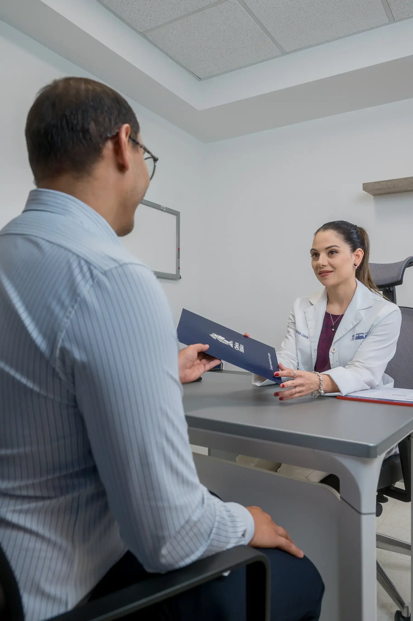 Médica especialista del Hospital Clínica Bíblica entregando una carpeta con resultados de exámenes a un paciente durante una consulta en su consultorio.