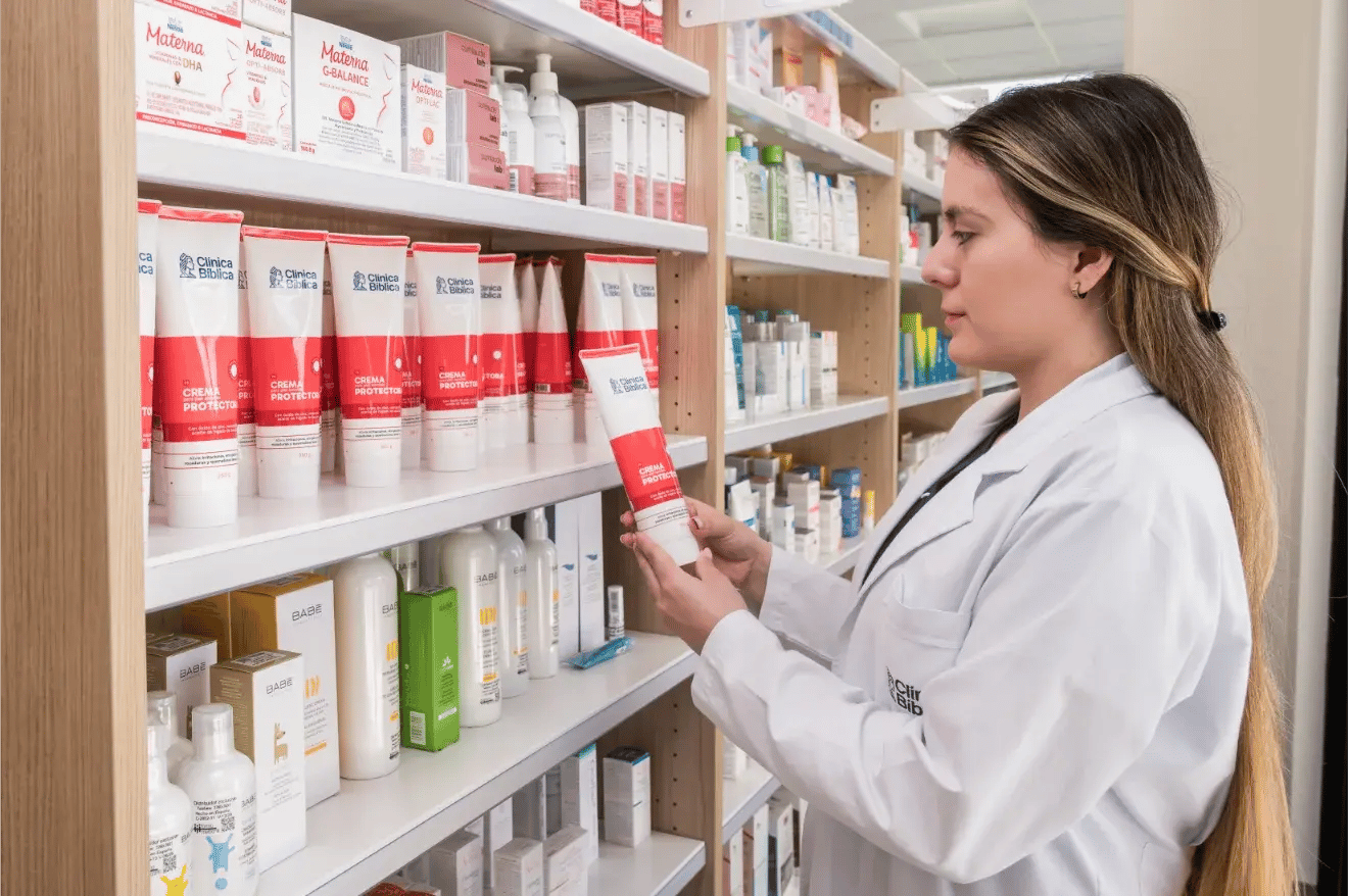 Farmacéutica del Hospital Clínica Bíblica revisando cremas dermatológicas y medicamentos en los estantes de la farmacia institucional.