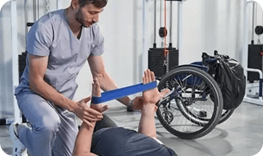 Fisioterapeuta del Hospital Clínica Bíblica asistiendo a un paciente en silla de ruedas durante ejercicios de rehabilitación de brazos con banda elástica.
