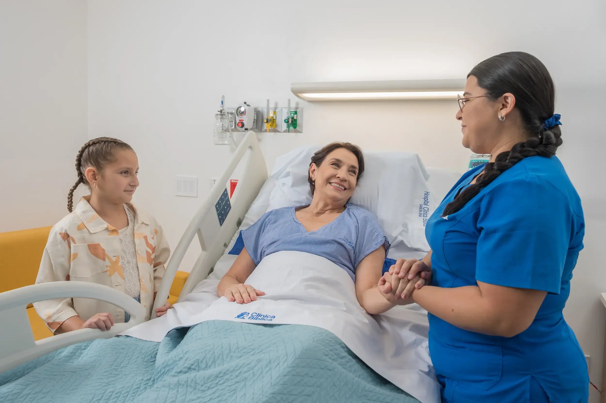 Enfermera del Hospital Clínica Bíblica conversando de forma empática con una paciente en su cama de hospitalización acompañada por una niña durante la visita familiar.