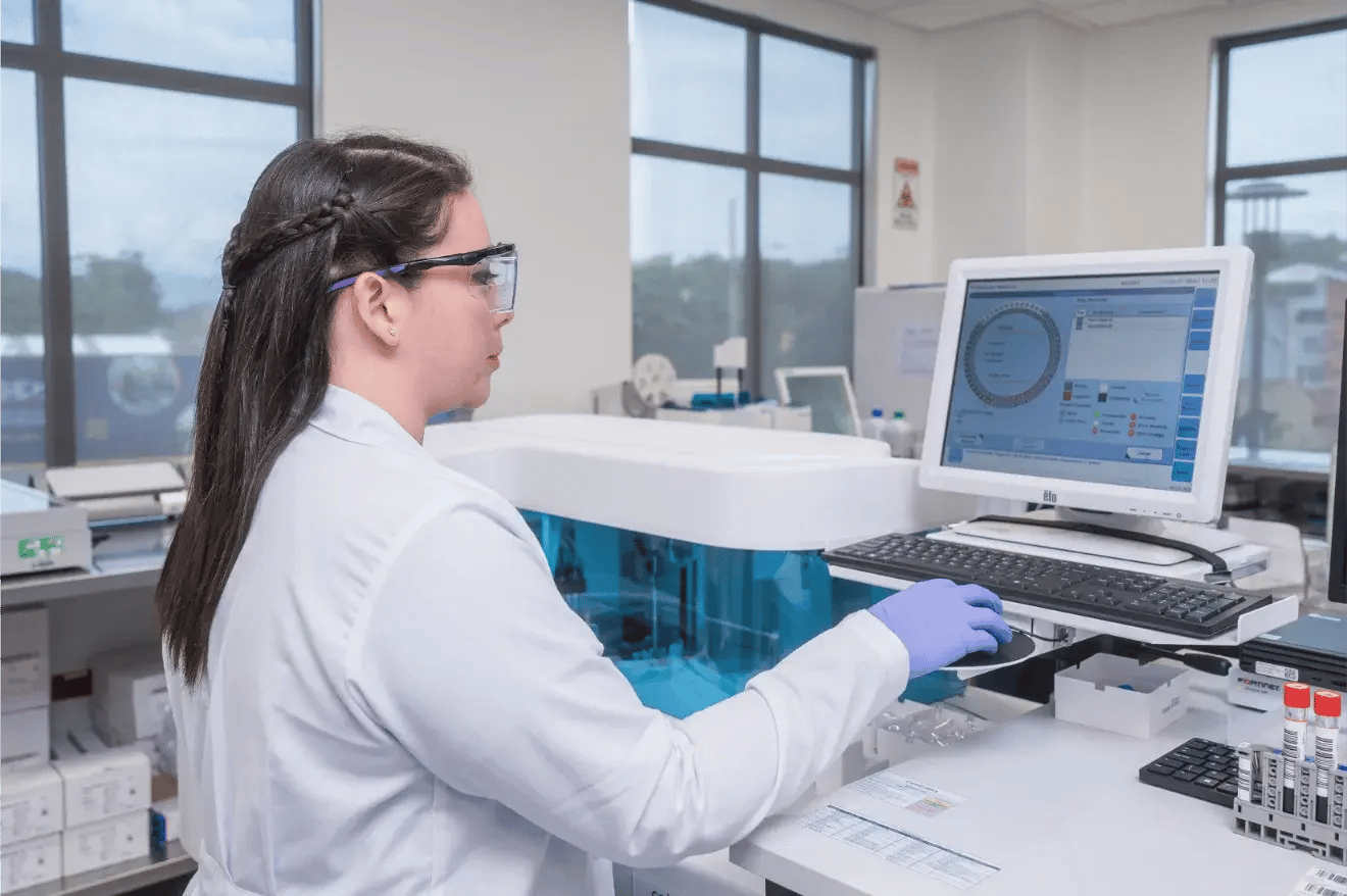 Especialista en laboratorio clínico del Hospital Clínica Bíblica procesando exámenes médicos mediante un analizador automatizado y software médico.
