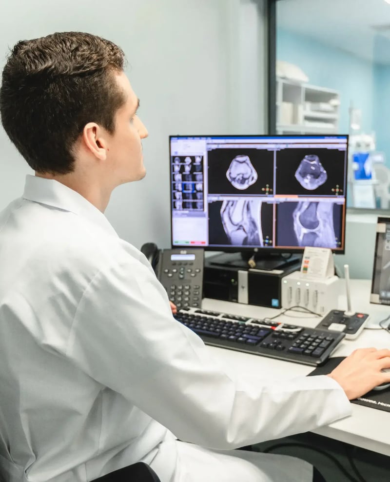Radiologist at Hospital Clinica Biblica analyzing a knee MRI scan on high-resolution monitors from the diagnostic imaging control room.