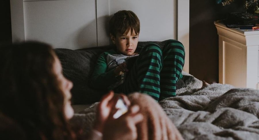 Un niño pequeño sentado en su cama por la noche, mirando con expresión seria y preocupada la pantalla de una tableta en una habitación con luz tenue.