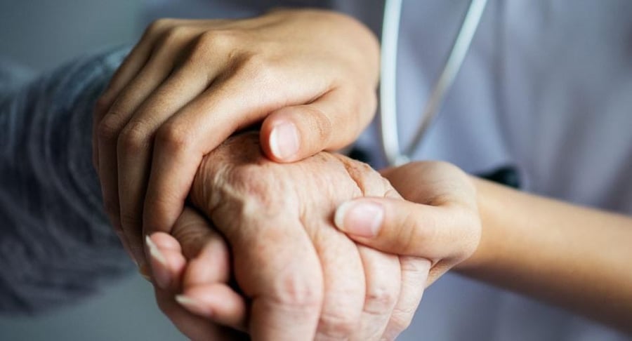 Primer plano de las manos de un profesional de la salud sosteniendo con ternura la mano de un adulto mayor, simbolizando apoyo y cuidado.