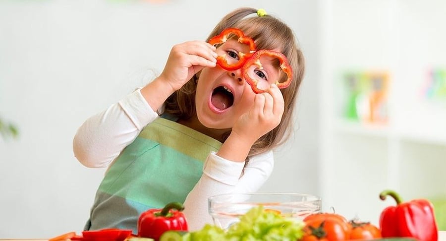 Niña pequeña con un delantal jugando alegremente en la cocina mientras se coloca dos rodajas de pimiento rojo sobre los ojos como si fueran anteojos.