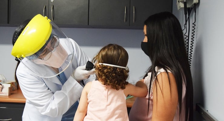 Doctora con equipo de protección revisando el oído de una niña pequeña acompañada por su madre en una consulta social.
