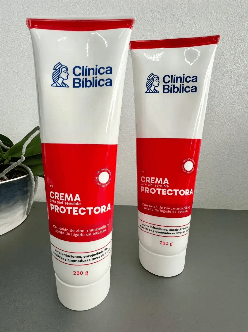 Dos tubos de crema protectora Clínica Bíblica sobre una mesa gris con empaque blanco y rojo