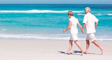 Pareja de adultos mayores caminando de la mano por una playa soleada, representando el disfrute de las vacaciones con un control adecuado de la diabetes.