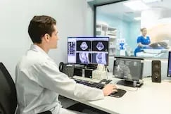 Radiólogo revisando imágenes de resonancia magnética en monitores, paciente en máquina de resonancia al fondo.