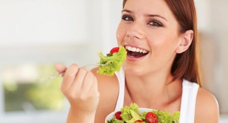 Mujer joven sonriente comiendo una ensalada fresca con lechuga y tomates cherry, representando una alimentación saludable y equilibrada.