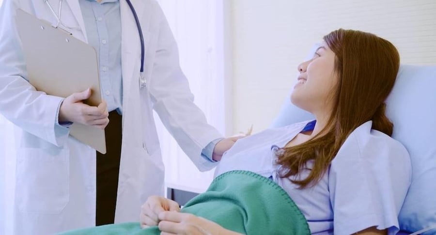 Médica especialista de Hospital Clínica Bíblica conversando con una paciente en cama sobre sus cuidados de recuperación en casa.