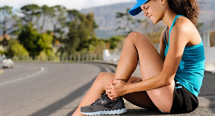 Mujer deportista sentada en la calle sujetando su tobillo con gesto de dolor durante actividad física.