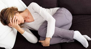 Mujer acostada en un sofá en posición fetal, cubriendo su rostro con una mano para expresar el malestar y la angustia causados por el dolor crónico.