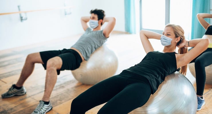 Personas realizando ejercicios abdominales sobre pelotas de yoga utilizando mascarillas quirúrgicas en un gimnasio.