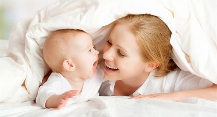 Madre y bebé acostados bajo una sábana mirándose y sonriendo