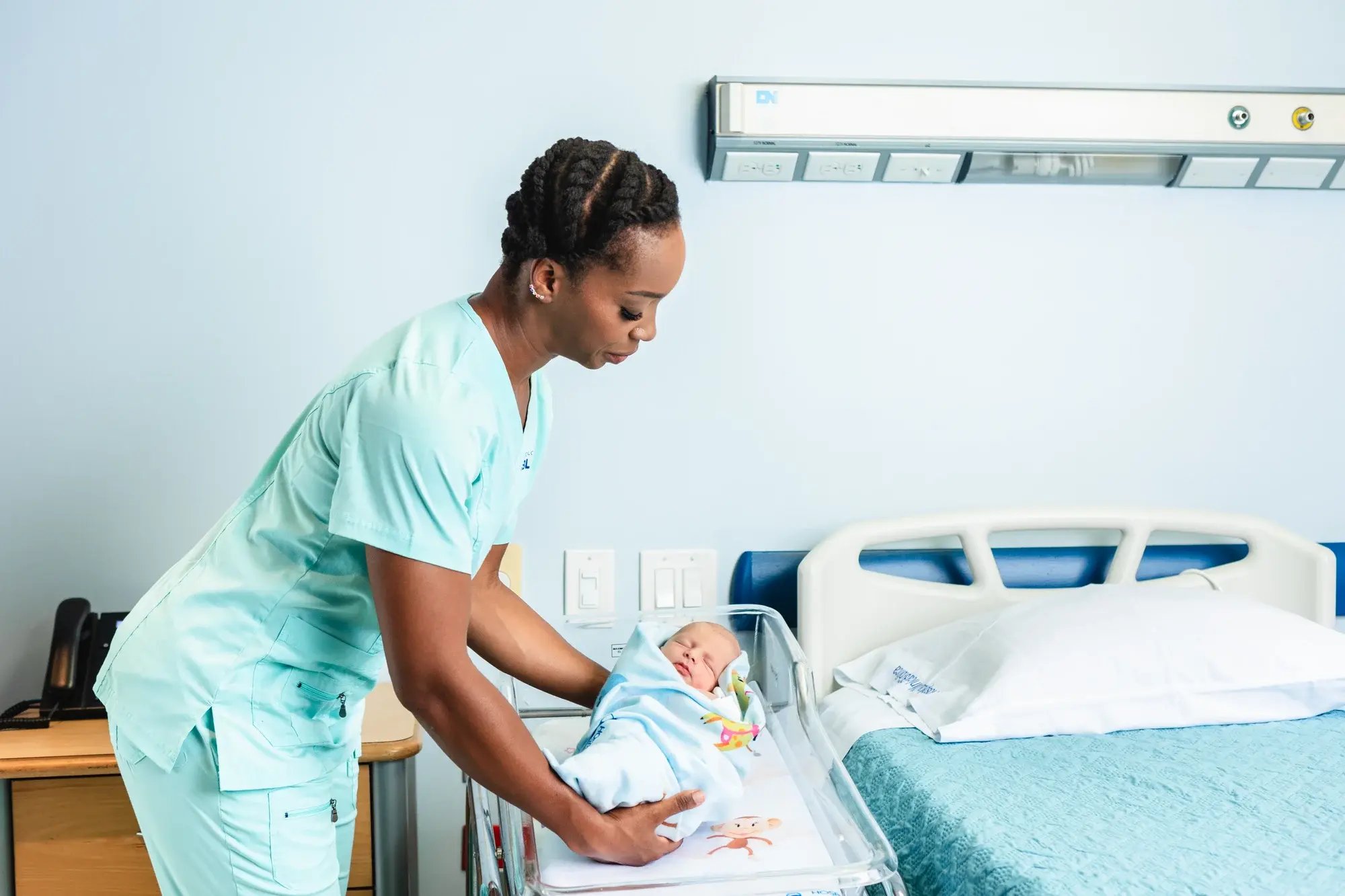 Enfermera acomodando a un recién nacido envuelto en una manta dentro de una cuna transparente en una habitación hospitalaria. Enfermera acomodando a un recién nacido envuelto en una manta dentro de una cuna transparente en una habitación hospitalaria.