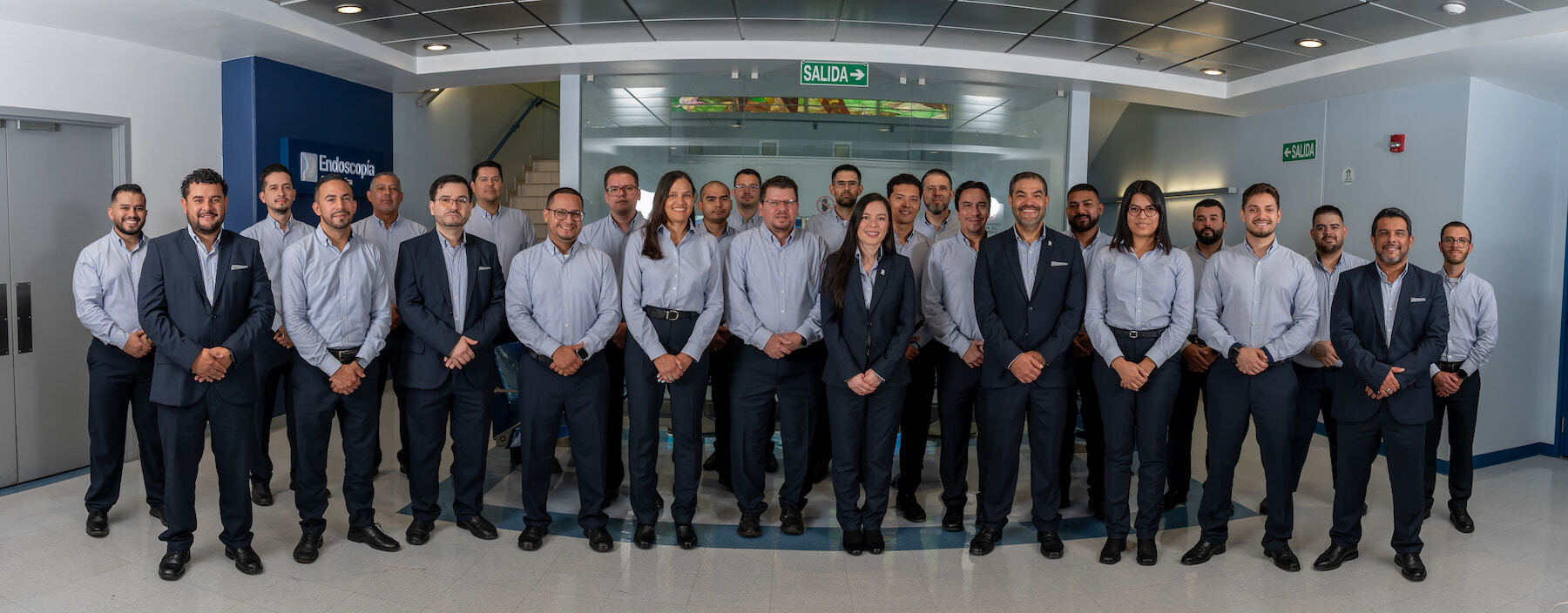 Fotografía grupal de un amplio equipo de colaboradores posando en el pasillo de un centro médico. Los profesionales visten uniformes corporativos formales, la mayoría con camisas de color celeste y pantalones oscuros, y algunos vistiendo trajes azul marino.