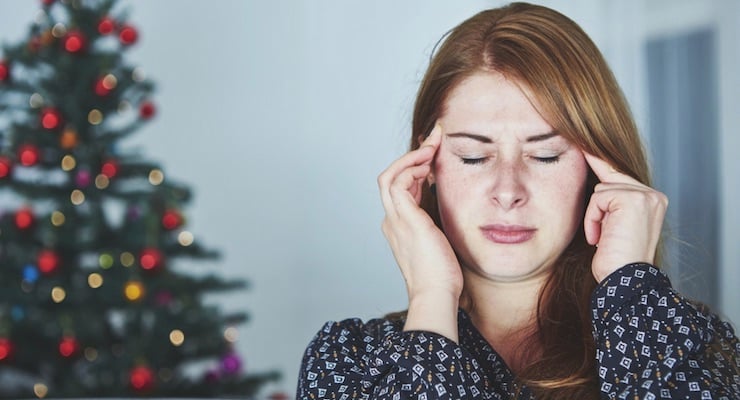 Mujer con expresión de estrés y dolor de cabeza frente a un árbol de Navidad desenfocado al fondo.