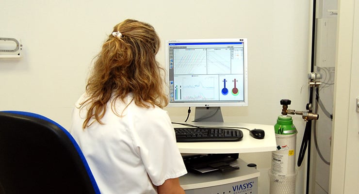 Profesional de la salud sentada frente a un equipo de neumología observando gráficos respiratorios en una pantalla de computadora.